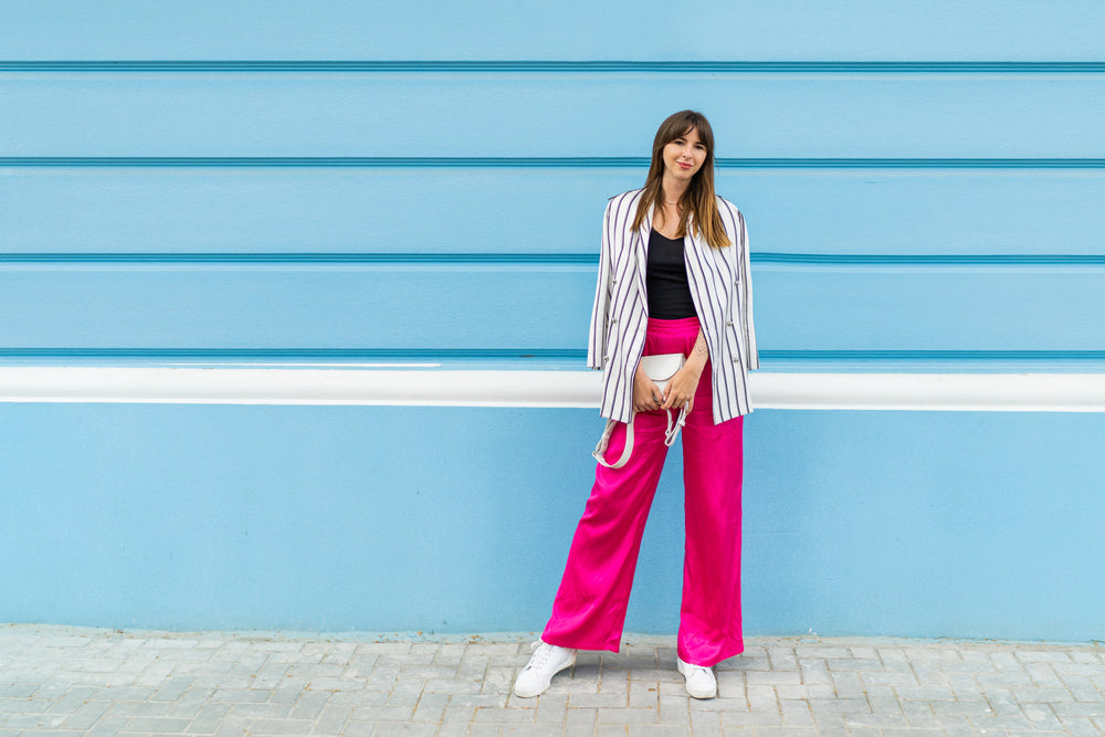 Mulher usando um look com calça rosa na rua, em frente a uma parede azul. 