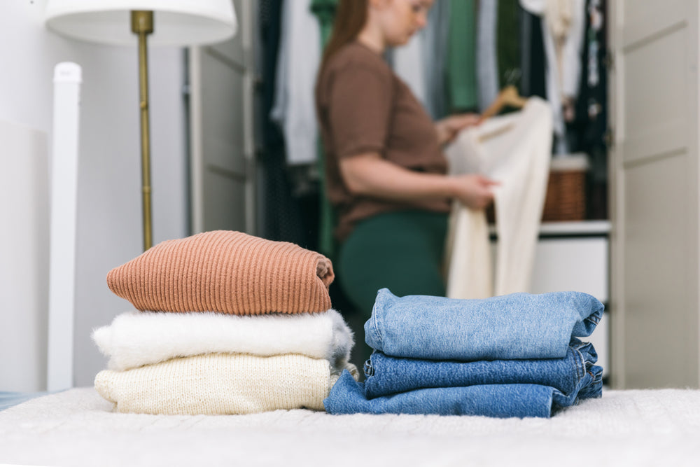 Em cima de uma cama, seis peças de roupas de diferentes tecidos dobradas. Ao fundo, uma mulher pendura uma peça de roupa num cabide, em frente a um guarda-roupa.