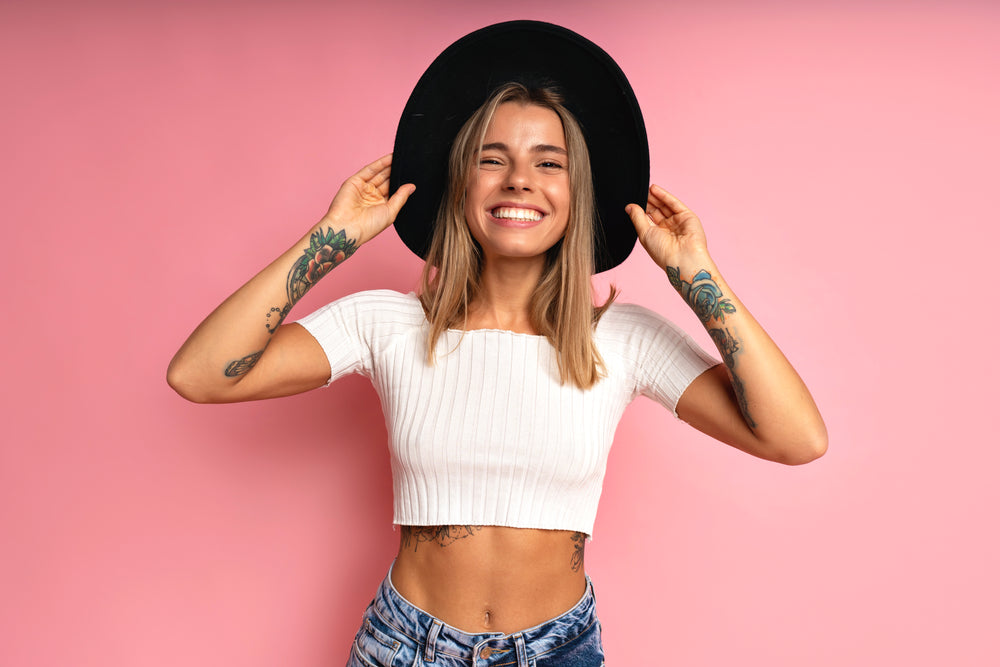ulher sorridente e estilosa posando no fundo de um estúdio rosa, vestindo top cropped branco e um chapéu fedora preto. Ela possui cabelos loiros e tatuagens nos braços.